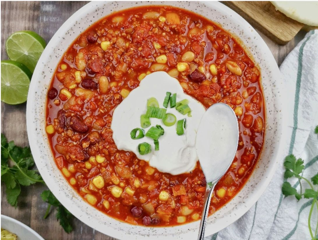 A top view of a bowl of chili topped with sour cream and chives on a table surrounded by a dish of brussel sprouts, a bowl of sour cream, and a wooden tray of sliced cheese.